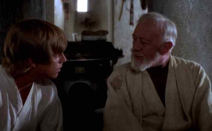 Ben Kenobi and Luke Skywalker are chatting in Ben's house, in this scene from Star Wars: A New Hope. Ben has a white beard and wears a beige tunic, Luke wears a white garment.