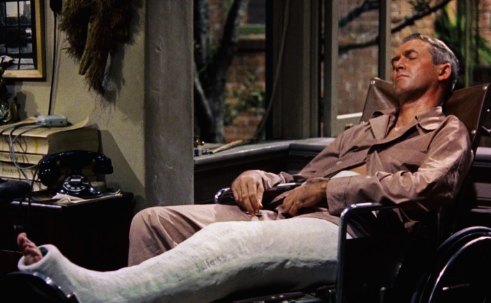 A scene from the film Rear Window. James Stewart sits in a chair with his leg in plaster.