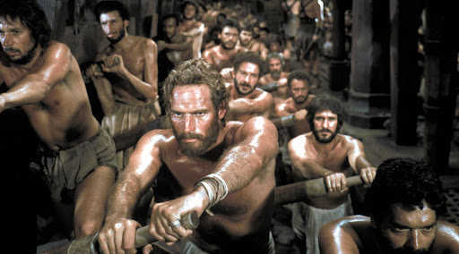 A scene from the film Ben-Hur. Charlton Heston is seated, rowing on a boat with other men.